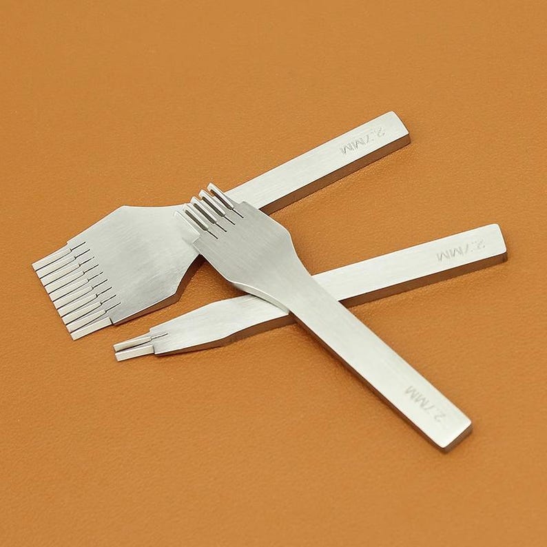 May include: Three silver leathercraft tools with different prong configurations are arranged on a brown surface. The tools include a multi-prong chisel, a four-prong chisel, and a two-prong chisel. Each tool is marked with "2.7MM".