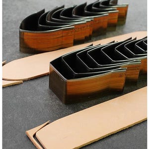 May include: Leatherworking tools, including a set of metal corner cutters and strips of tan leather. The cutters are arranged in a row, with a wooden-like finish on the sides. The leather strips are light brown and appear to be freshly cut.