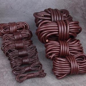 Può includere: Diversi fasci di cordoncino in pelle marrone scuro sono disposti su una superficie grigia. I cordoncini sono strettamente avvolti e legati, mostrando un colore ricco e profondo. Le diverse dimensioni suggeriscono lunghezze variabili per lavori artigianali.