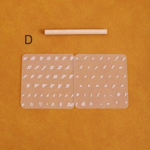 May include: A clear, flexible alphabet and number stencil set with a wooden dowel. The stencil includes uppercase and lowercase letters, numbers, and symbols. The letter "D" is visible in the upper left corner. The background is a solid orange color.