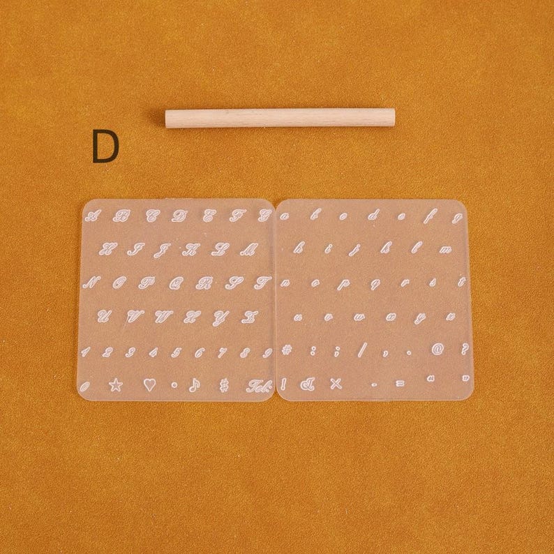 May include: A clear, flexible alphabet and number stencil set with a wooden dowel. The stencil includes uppercase and lowercase letters, numbers, and symbols. The letter "D" is visible in the upper left corner. The background is a solid orange color.