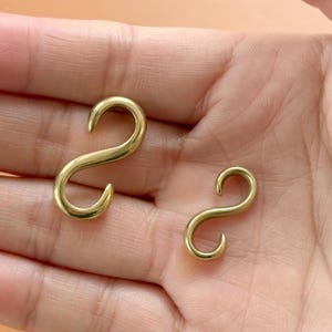 May include: Two brass-colored S-hooks, each with a curved, S-shaped design. The hooks are displayed against a neutral background, highlighting their metallic sheen. These hooks are suitable for various hanging and organizational purposes.