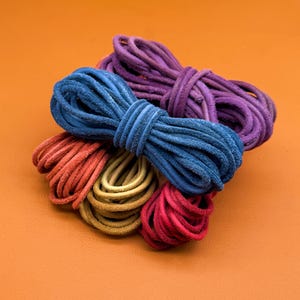 Può includere: Una collezione di cinque cordini in pelle avvolti in vari colori, tra cui viola, blu, rosso, giallo e rosa. Ogni cordino è avvolto in un fascio stretto, mostrando una consistenza morbida e opaca e un profilo arrotondato.