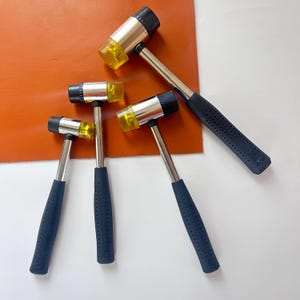 Pode incluir: Uma coleção de cinco martelos de borracha. Cada martelo apresenta uma cabeça de impacto amarela, um eixo de metal prateado e uma pega azul marinho texturizada. Os martelos estão dispostos sobre uma superfície branca com um quadrado de couro castanho no fundo.