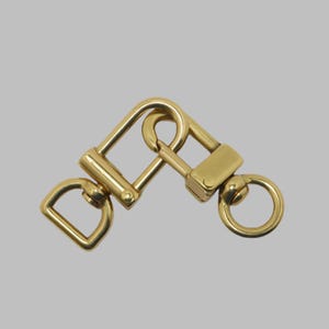 May include: A gold-colored swivel clasp with a D-ring, a square loop, and a circular ring. The metal hardware is designed to connect two straps or other items. The clasp is made of a shiny, durable metal.