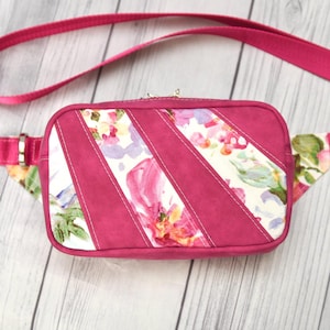 Op de afbeelding: Een fuchsia kleurige heuptas met een bloemenpatroon en een bijpassende verstelbare riem. De tas heeft een rechthoekige vorm met diagonale panelen van bloemenstof en effen fuchsia materiaal. Het heeft een ritssluiting en een zilverkleurige sluiting.