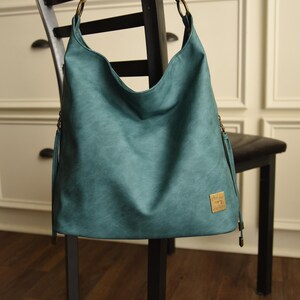 Op de afbeelding: Teal schoudertas met een gebogen bovenkant en een getextureerde uitstraling. De tas heeft een goudkleurig metalen label en bijpassende hardware op de bandjes. De tas hangt aan een zwarte stoel.
