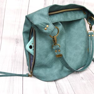 Op de afbeelding: Petrolkleurige hobo-tas met een ritsvak dat een lichtblauwe telefoonhoes onthult. De tas heeft een bovenrits, messingkleurige hardware en een afneembare riem. De tas is gemaakt van een textuurrijk materiaal.