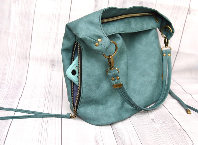 Op de afbeelding: Petrolkleurige hobo-tas met een ritsvak dat een lichtblauwe telefoonhoes onthult. De tas heeft een bovenrits, messingkleurige hardware en een afneembare riem. De tas is gemaakt van een textuurrijk materiaal.