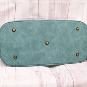 Op de afbeelding: Onderkant van een teal handtas met een getextureerde afwerking. De tas heeft een afgeronde vorm met messingkleurige voetjes en hardware. De ritstrekker en de bandjes zijn ook messingkleurig.