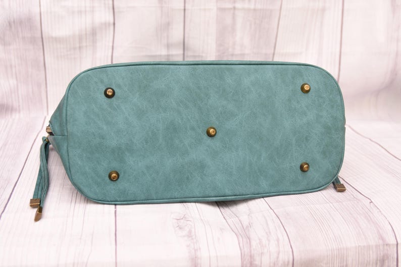 Op de afbeelding: Onderkant van een teal handtas met een getextureerde afwerking. De tas heeft een afgeronde vorm met messingkleurige voetjes en hardware. De ritstrekker en de bandjes zijn ook messingkleurig.
