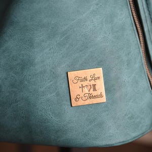 Op de afbeelding: Close-up van een teal-kleurige tas met een getextureerd oppervlak. Een klein, vierkant, bronzen label is bevestigd, gegraveerd met de woorden "Faith Love & Threads", een kruis, een hart en een klos garen.