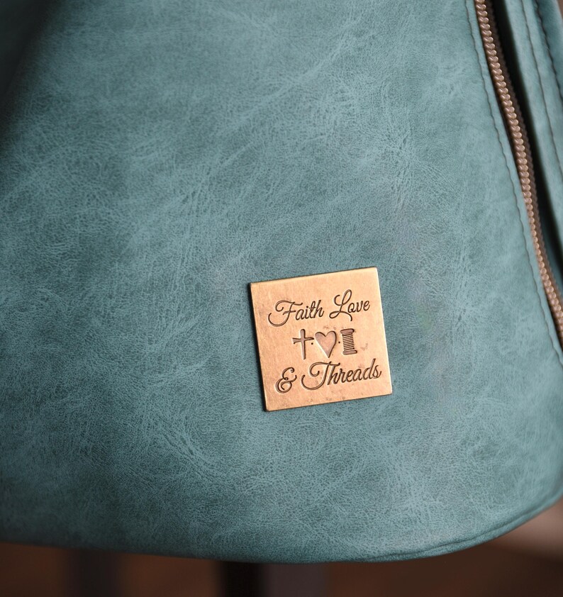 Op de afbeelding: Close-up van een teal-kleurige tas met een getextureerd oppervlak. Een klein, vierkant, bronzen label is bevestigd, gegraveerd met de woorden "Faith Love & Threads", een kruis, een hart en een klos garen.
