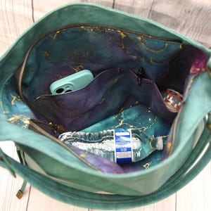 Op de afbeelding: Teal handtas met een ruim interieur met meerdere vakken. De binnenvoering toont een hemels patroon in tinten blauw, paars en goud. Een waterfles en een telefoon zijn zichtbaar in de tas. De tas heeft een ritssluiting en een schouderriem.
