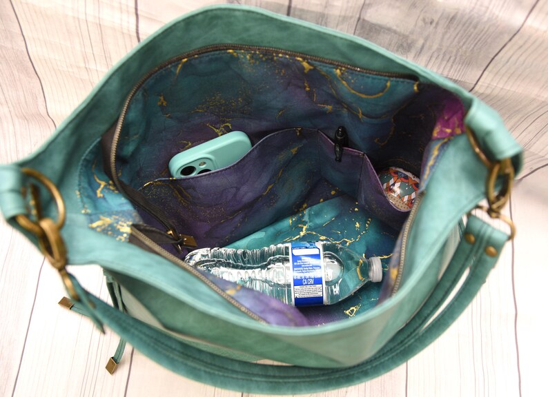 Op de afbeelding: Teal handtas met een ruim interieur met meerdere vakken. De binnenvoering toont een hemels patroon in tinten blauw, paars en goud. Een waterfles en een telefoon zijn zichtbaar in de tas. De tas heeft een ritssluiting en een schouderriem.