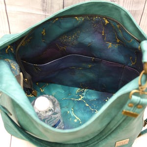 Op de afbeelding: Teal handtas met een ruim interieur. De binnenkant heeft een teal, paarse en gouden gemarmerde stoffen voering, met meerdere vakken en een vak met ritssluiting. Een waterfles is zichtbaar in de tas.