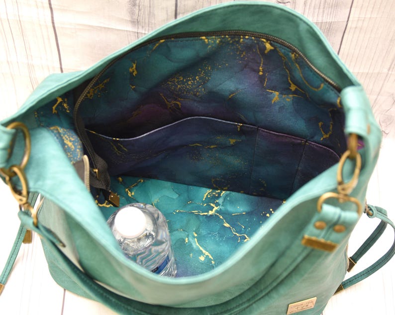 Op de afbeelding: Teal handtas met een ruim interieur. De binnenkant heeft een teal, paarse en gouden gemarmerde stoffen voering, met meerdere vakken en een vak met ritssluiting. Een waterfles is zichtbaar in de tas.