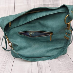 Op de afbeelding: Teal schoudertas met een getextureerde afwerking. De tas heeft een voorvak met ritssluiting en een hoofdvak met ritssluiting. Het heeft goudkleurige hardware, inclusief ringen en ritsen, en een lange riem.