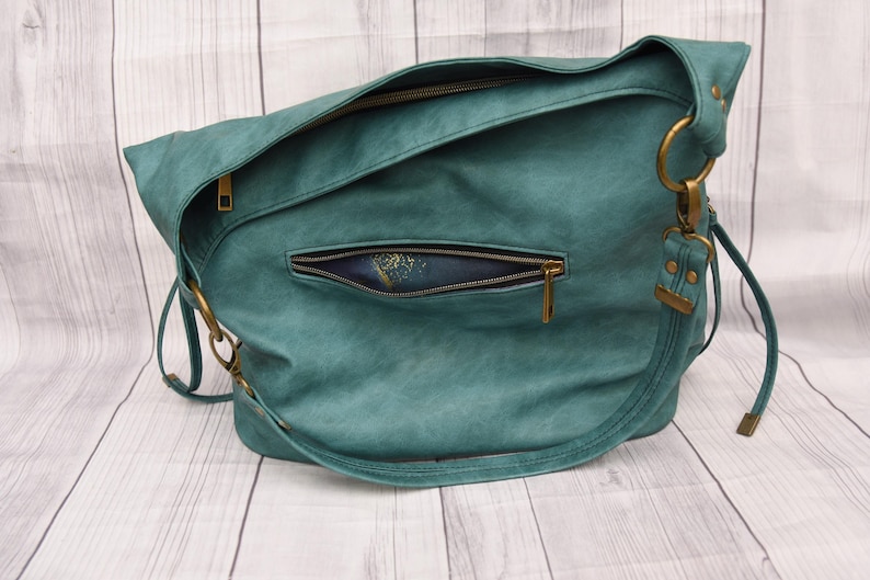Op de afbeelding: Teal schoudertas met een getextureerde afwerking. De tas heeft een voorvak met ritssluiting en een hoofdvak met ritssluiting. Het heeft goudkleurige hardware, inclusief ringen en ritsen, en een lange riem.
