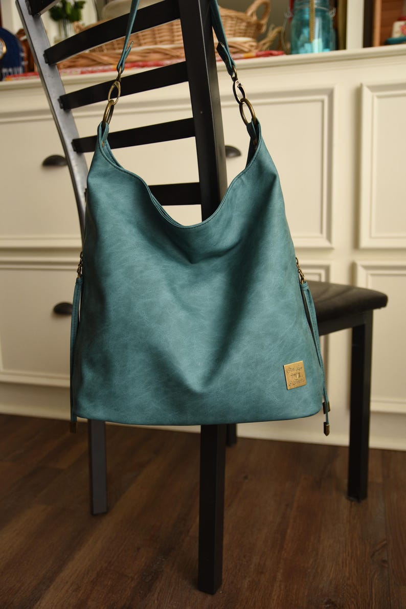 Op de afbeelding: Teal schoudertas met een gebogen bovenkant en een getextureerde uitstraling. De tas heeft een goudkleurig metalen label en bijpassende hardware op de bandjes. De tas hangt aan een zwarte stoel.
