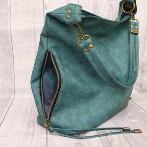 Op de afbeelding: Teal schoudertas met een zijvak met ritssluiting en een lange riem. De tas heeft goudkleurige hardware, inclusief klinknagels, sluitingen en ritstrekkers. De binnenvoering is zichtbaar door de open rits.