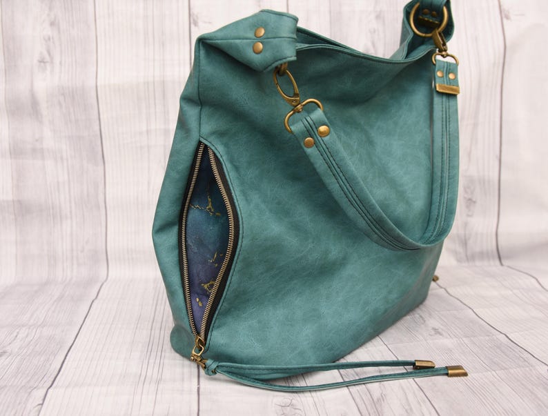 Op de afbeelding: Teal schoudertas met een zijvak met ritssluiting en een lange riem. De tas heeft goudkleurige hardware, inclusief klinknagels, sluitingen en ritstrekkers. De binnenvoering is zichtbaar door de open rits.