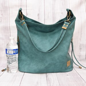 Op de afbeelding: Teal-kleurige hobo-tas met een schouderriem en goudkleurige hardware. De tas heeft een getextureerd uiterlijk en een klein rechthoekig label. Er staat een waterfles naast de tas. De tas is ongeveer 36 cm breed.