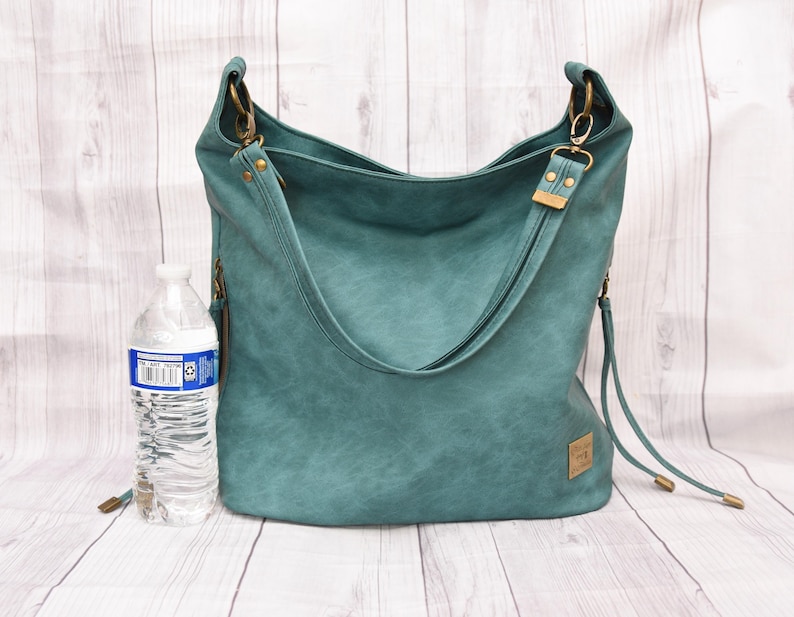 Op de afbeelding: Teal-kleurige hobo-tas met een schouderriem en goudkleurige hardware. De tas heeft een getextureerd uiterlijk en een klein rechthoekig label. Er staat een waterfles naast de tas. De tas is ongeveer 36 cm breed.