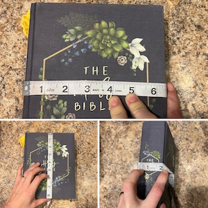 May include: A dark grey book titled "The Holy Bible" with floral and geometric designs. The book is being measured with a tape measure, showing a width of approximately 10 cm and a height of approximately 20 cm.