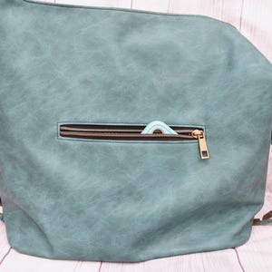 Op de afbeelding: Een teal rugzak met een voorvak met ritssluiting. De rits is bronskleurig en een klein, lichtblauw item is gedeeltelijk zichtbaar in de zak. De tas heeft verstelbare bandjes en een getextureerde uitstraling.