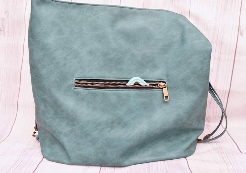 Op de afbeelding: Een teal rugzak met een voorvak met ritssluiting. De rits is bronskleurig en een klein, lichtblauw item is gedeeltelijk zichtbaar in de zak. De tas heeft verstelbare bandjes en een getextureerde uitstraling.