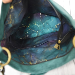 Op de afbeelding: Open zicht op een teal handtas met gouden details. De binnenkant heeft een gemarmerd patroon in tinten van teal, blauw, paars en goud. De tas heeft een zwarte band en goudkleurige ringen.