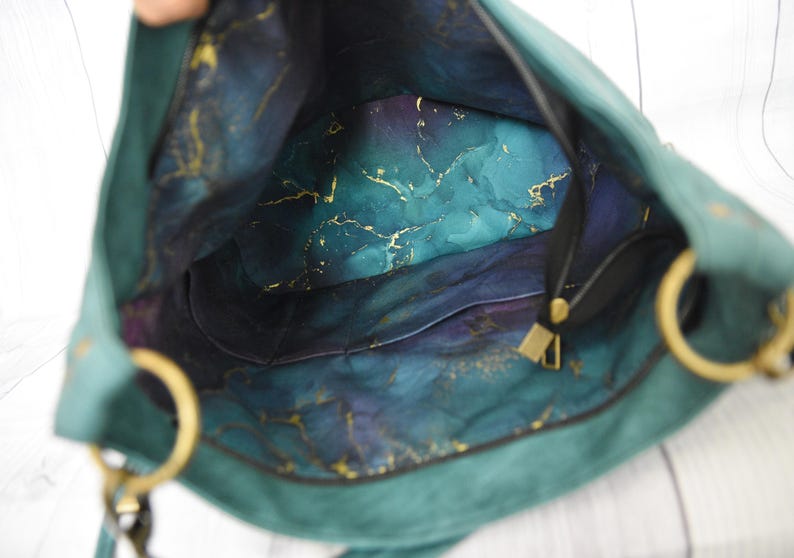 Op de afbeelding: Open zicht op een teal handtas met gouden details. De binnenkant heeft een gemarmerd patroon in tinten van teal, blauw, paars en goud. De tas heeft een zwarte band en goudkleurige ringen.
