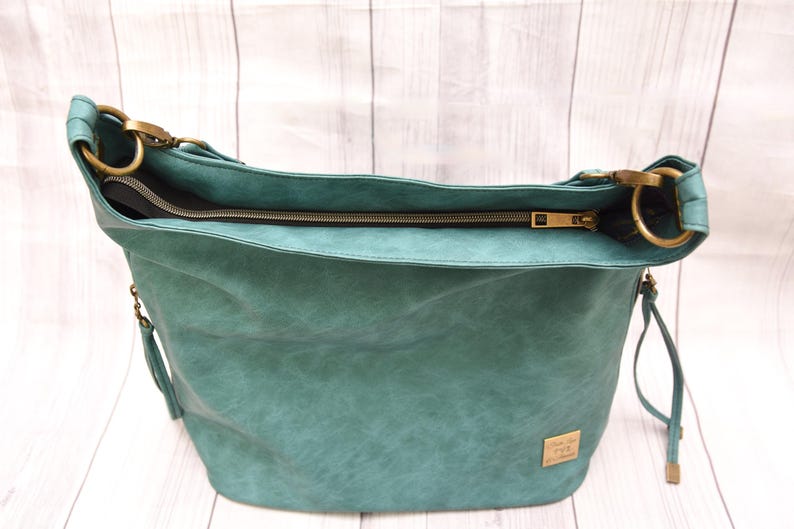 Op de afbeelding: Teal schoudertas met een getextureerde afwerking. De tas heeft een ritssluiting aan de bovenkant, goudkleurige hardware en een klein rechthoekig label. De tas heeft een nonchalant ontwerp en een lange riem.