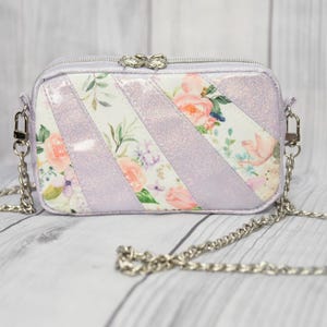 Op de afbeelding: Kleine, rechthoekige crossbodytas met een zilveren kettingriem. De tas heeft een lavendelkleurige buitenkant met diagonale strepen van bloemenstof. De rits heeft een decoratieve zilveren trekker.