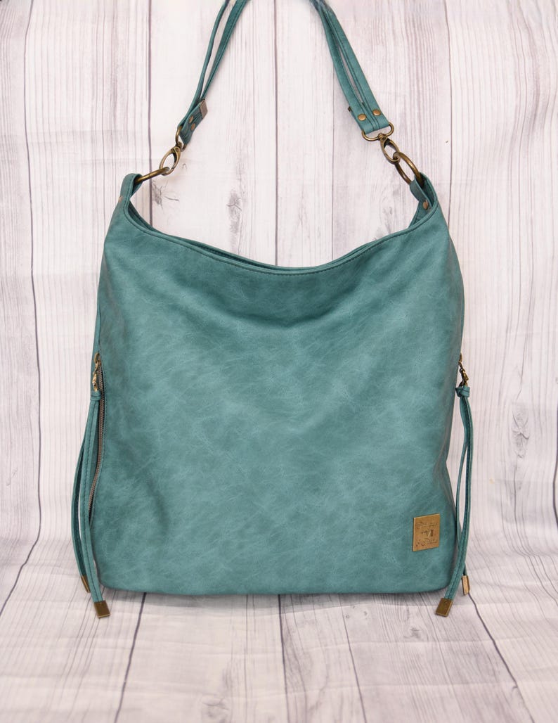 Op de afbeelding: Teal schoudertas met een distressed, getextureerde uitstraling. De tas heeft lange banden, metalen hardware en een klein rechthoekig label. De tas heeft een groot, open ontwerp en is gemaakt van een zacht materiaal.