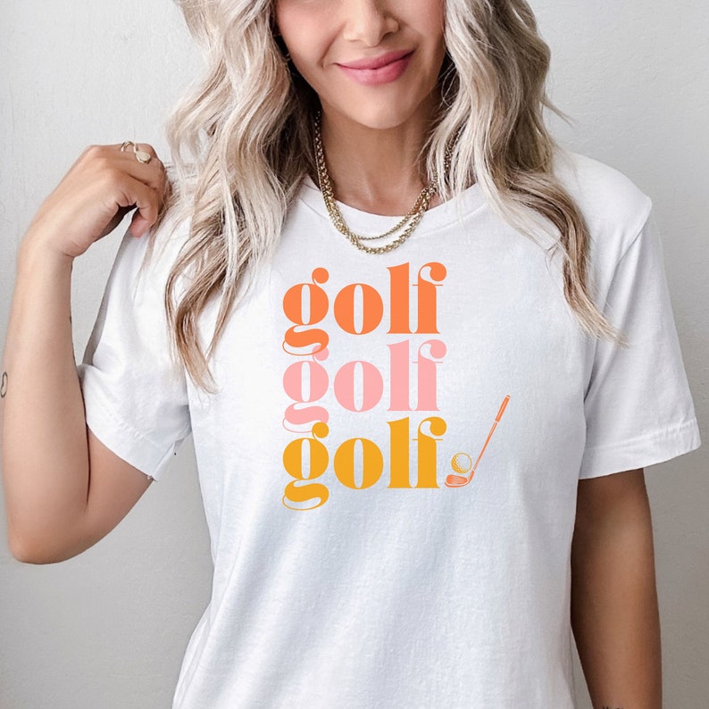 May include: White t-shirt with the words "golf golf golf" printed in orange, pink, and yellow. A golf club and ball are also printed on the shirt.