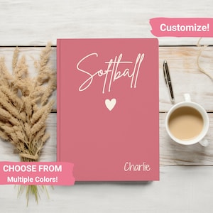 May include: A pink notebook with the word "Softball" written in white cursive script. There is a small white heart below the word. The name "Charlie" is written in white at the bottom of the notebook. The notebook is on a wooden surface with a cup of coffee and a pen.