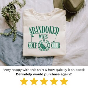 May include: A beige t-shirt with a green graphic that says "Abandoned Wives Golf Club". The graphic includes a golf club, a golf ball, and a flag.