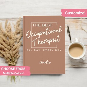 May include: A brown notebook with the text "THE. BEST. Occupational Therapist ALL DAY. EVERY DAY." and the name "charlie" written below. The notebook is on a wooden surface with a cup of coffee and a pen.