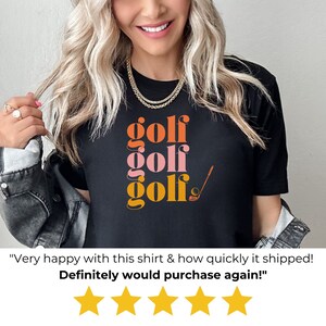 May include: Black t-shirt with the word "golf" repeated three times in stacked, colorful text. The word "golf" is in orange, pink, and yellow. A golf club and ball are to the right of the text.