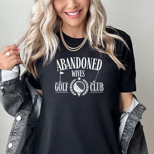 May include: Black t-shirt with white text that reads "Abandoned Wives Golf Club". The text is in a vintage style font and includes a golf ball and club graphic.