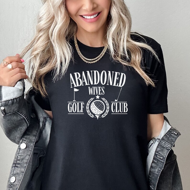May include: Black t-shirt with white text that reads "Abandoned Wives Golf Club". The text is in a vintage style font and includes a golf ball and club graphic.