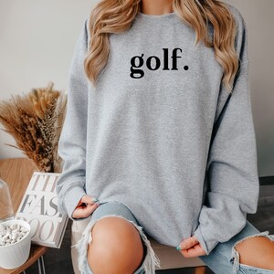 Woman wearing light gray “golf.” sweatshirt, sitting comfortably — relaxed and casual golf lover sweatshirt.