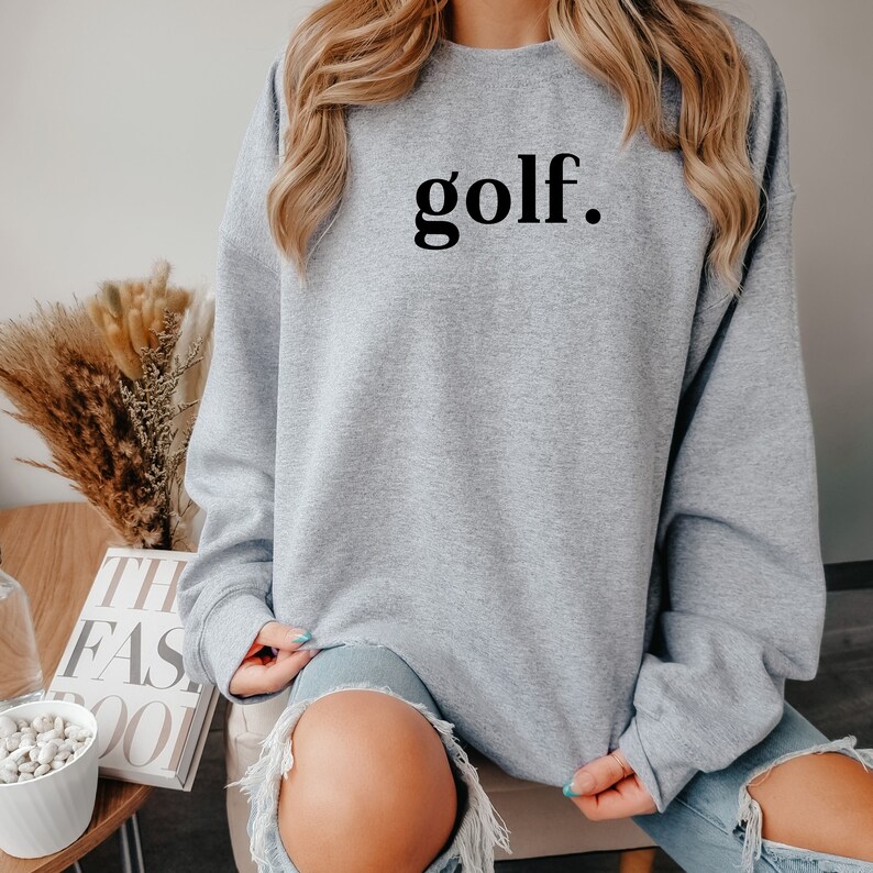 Woman wearing light gray “golf.” sweatshirt, sitting comfortably — relaxed and casual golf lover sweatshirt.