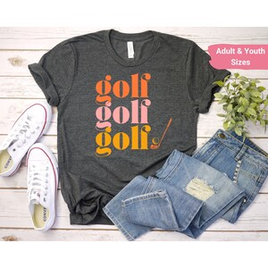 May include: A dark gray t-shirt with the words "golf golf golf" in orange, pink, and yellow stacked on top of each other. A golf club and ball are next to the word "golf".