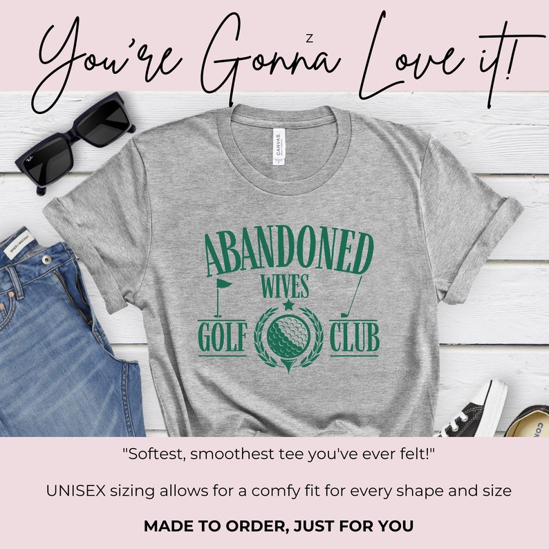 May include: Grey t-shirt with green text that reads "Abandoned Wives Golf Club". The text is surrounded by a green wreath with a golf ball in the centre. The shirt is on a wooden surface with a pair of sunglasses and a pair of jeans.
