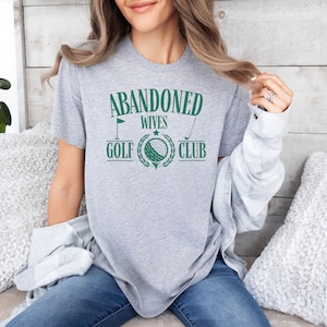 May include: A grey t-shirt with the text "Abandoned Wives Golf Club" in green lettering. The text is surrounded by a green wreath with a golf ball in the centre.