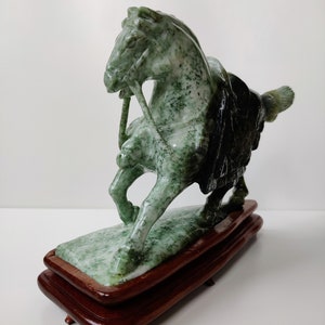 May include: A green jade horse sculpture on a wooden base. The horse is standing on its hind legs with its head turned to the side. The sculpture is detailed with intricate carvings.