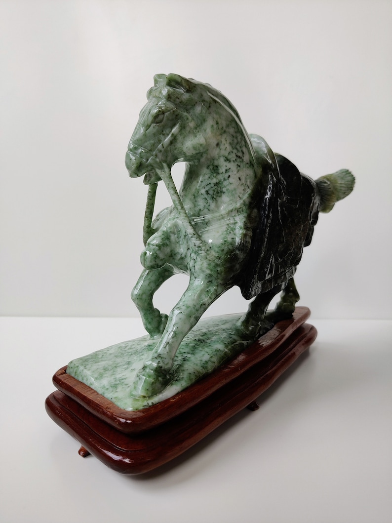 May include: A green jade horse sculpture on a wooden base. The horse is standing on its hind legs with its head turned to the side. The sculpture is detailed with intricate carvings.